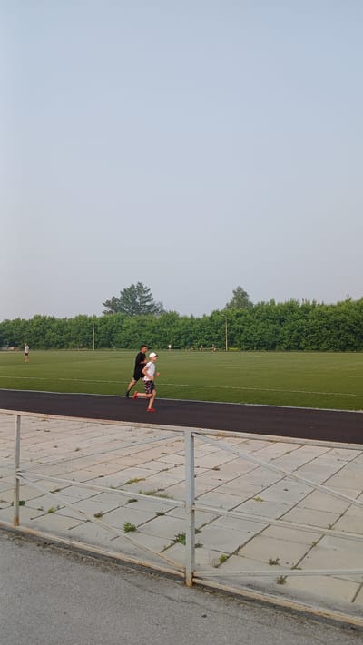 The image depicts an outdoor athletic track in Kolyvan, Russia, on a hazy day. Two individuals are running on the track. In the foreground, a person wearing a black t-shirt and shorts is running, accompanied by a younger person in a white t-shirt and patterned shorts. Behind them, other individuals can be seen running and walking on the green field. A line of trees borders the field, and a light-colored sky with visible haze occupies the upper portion of the frame. A metal fence and a concrete path are also visible in the foreground.