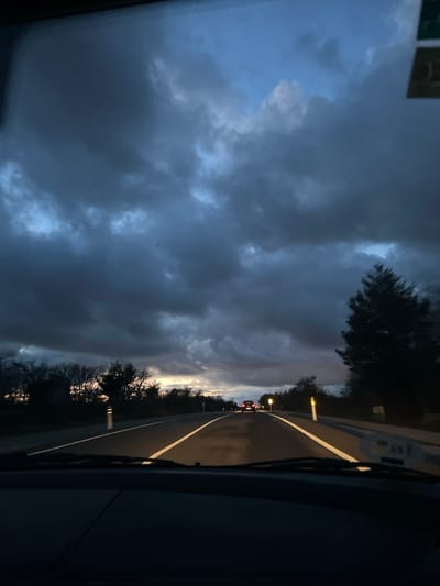 The image displays a forward view from inside a vehicle on a paved, two-lane road within or near Real Sitio de San Ildefonso, Spain, at twilight. The sky is predominantly covered by heavy, dark grey and bluish-grey clouds, with visible patches of lighter blue sky in the upper areas. A muted band of yellow-orange light is present on the horizon, primarily to the left, indicating sunrise or sunset. The road features clear solid white lane markings and exhibits a slightly reflective surface. In the distance, several vehicles are observed traveling in the same direction, identified by their illuminated red taillights. The left side of the road is bordered by a line of dark trees, possibly bare, and two white-and-black striped bollards. On the right side, a large, dark conifer tree stands near the road, accompanied by another white-and-black striped bollard. The lower portion of the image shows the dark dashboard and the base of the windshield of the originating vehicle.