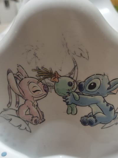 The scene is situated in Velichkovo, Bulgaria. The image presents a close-up view of a smooth, white, curved surface, likely ceramic or plastic, adorned with a multi-color graphic. The graphic depicts three stylized animated characters. On the left, a pink character identified as Angel (Experiment 624) is depicted seated, facing right, with closed eyes and a contented facial expression. On the right, a blue character identified as Stitch (Experiment 626) is shown, oriented towards Angel, holding and presenting a small, green, humanoid doll. This doll features black button eyes, visible stitching across its body, and a top-knot of yellow fibers secured by a red-orange bow. Both animated characters are rendered with a sketch-like aesthetic, featuring distinct outlines and subtle color shading. Faint, incomplete line drawings of tropical foliage, specifically palm tree fronds, are visible in the background above the characters. The white surface exhibits minimal superficial wear.