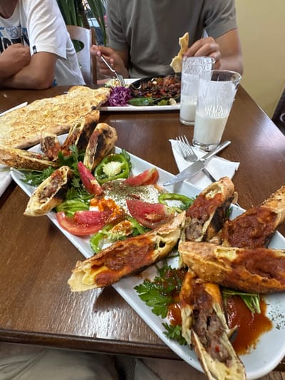 The image depicts a table laden with food, suggesting a meal in a restaurant setting, likely in Gelemenovo, Bulgaria. In the foreground, a white rectangular platter overflows with several pieces of what appears to be Turkish-style pizza or borek, filled with meat and topped with a red sauce. Accompanying the pizza are sliced tomatoes, green peppers, and fresh parsley. To the left of the platter, a large piece of flatbread is visible. In the background, two glasses filled with a white beverage, possibly ayran, are placed on the table. The hands of at least two individuals are visible, one holding a fork and interacting with a dish in the background, and another holding a piece of bread. The table is made of wood, and the overall ambiance suggests a casual dining experience.
