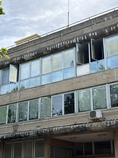 The image displays the facade of a multi-story building, shot from a low, upward-looking angle, with the structure tilted diagonally across the frame. The building is constructed of raw concrete, showing significant weathering, discoloration, and dark streaks, possibly from water runoff. White, crust-like deposits are visible along the horizontal concrete beams.

The facade is dominated by numerous rectangular windows, arranged in a grid-like pattern. Most windows have aluminum or white frames. Many are dirty or aged, with some showing internal reflections of trees and the cloudy sky, while others have a frosted or opaque appearance, possibly due to old film or grime. Several windows are open, tilted inwards from the top, revealing dark interiors.

On the upper right side of the facade, an outdoor air conditioning unit is mounted on a metal bracket. Below it, a section of the building's side wall, perpendicular to the main facade, is visible. This section is painted a lighter color and features two decorative, symmetrical fixtures, possibly sconces or small architectural embellishments.

To the left of the building, a bright, cloudy sky fills the background. A thin metal railing runs horizontally along the edge of the concrete structure, with wires or cables strung below it. Green foliage is visible in the bottom left corner and reflected in several windows, suggesting nearby trees. The overall impression is one of an aged urban structure, likely in Portoferraio, Italy, given the location context. No people or legible text are visible. The time of day is clearly daylight, under an overcast sky.