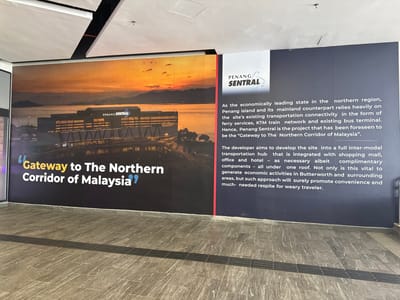 A large wall mural showcases the Penang Sentral transportation hub. One side features a sunset view with the building prominently displayed and the phrase "Gateway to The Northern Corridor of Malaysia." The other side provides a description of the project, highlighting its role in connecting Penang Island with the mainland through ferry services, train networks, and bus terminals. It also mentions the development's aim to be an inter-modal transportation hub with shopping, office, and hotel components to boost the economy and offer convenience to travelers.