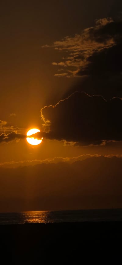 The image depicts a sunset over the sea, casting a warm, orange glow across the sky and water. The sun, a bright, luminous orb, is partially obscured by dark, dramatic clouds, creating a striking contrast between light and shadow. The clouds, tinged with the sunset's hues, drift across the sky, some appearing dense and heavy, while others are wispy and ethereal. The sea below reflects the fiery colors of the sky, with a shimmering path of light stretching across the surface. The foreground is a dark, silhouetted expanse, possibly a beach or landmass, grounding the scene. The overall atmosphere is one of dramatic beauty and natural splendor. This scene does not show any indication of being in Boz Beshik, Kyrgyzstan, as it depicts a coastal environment.