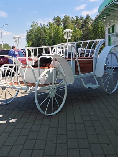 In a paved outdoor area, likely near a roadside service stop given the presence of vehicles and a building with a green roof, a decorative white carriage is the primary subject. The carriage, designed to resemble a horse-drawn vehicle, features large, spoked wheels, a wooden interior, and ornamental railings. A black helmet rests on the carriage seat, suggesting it might be part of a display or recreational offering.

Two individuals are visible. One man, wearing a blue shirt, stands near a red car, appearing to be interacting with it. Another person, dressed in dark clothing, is partially visible within the carriage structure. The scene is illuminated by bright daylight, indicating it is daytime. The sky is blue with scattered white clouds, and a line of lush green trees forms a backdrop, suggesting a pleasant, possibly warm weather day.

The setting appears to be a public area, possibly a rest stop or a park, given the combination of paved walkways, parked cars, and trees. The overall atmosphere is casual and relaxed. The image does not contain any clearly legible text. The location context provided is Лапіцкі сельскі Савет, Belarus.