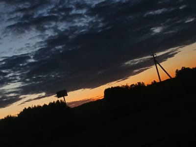 The image depicts a twilight sky over a rural landscape, likely in Воскресенское, Russia. Dark, heavy clouds dominate the upper portion of the frame, obscuring much of the sky. Below the clouds, a sliver of orange and yellow sunset is visible near the horizon, casting a warm glow. The silhouette of a dense line of trees runs horizontally across the mid-ground, with their dark forms sharply defined against the fading light. A solitary, angular utility pole with crossbars stands prominently to the right of the frame, also silhouetted. To the left, a dark, rectangular sign on a pole emerges from the tree line. The foreground is almost entirely black, suggesting deep shadow or darkness. The overall impression is one of approaching night in a natural or semi-rural setting.