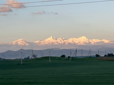 The image depicts a rural landscape in Nartan, Russia, under a pale sky with scattered clouds. A vast green field dominates the foreground. In the mid-ground, rolling hills are interspersed with utility poles. Beyond the hills, a mountain range with snow-capped peaks is visible. The lighting suggests either early morning or late evening. No people or animals are visible.