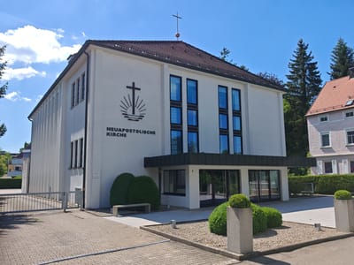 The image shows the exterior of the Neuapostolische Kirche in Memmingen, Germany. The building is white with a brown tiled roof and a cross on top. It has several large windows and a covered entrance with glass doors. In front of the building, there is a paved area with a metal gate, a stone bench, and several manicured bushes. To the right of the church, a portion of a pink-colored building with white shutters is visible. The sky is blue with scattered clouds, and tall evergreen trees are present in the background. The scene is well-lit, suggesting it was taken on a sunny day.