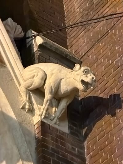 A gargoyle statue perches on a ledge against a brick building. It is light-colored, with its mouth open in a possible roar or yawn. It appears to be on a building in Osijek, Croatia. The statue's shadow is cast on the brickwork behind it. Two dark wires angle downward on the left side. The background shows a diagonally laid brick pattern. The bottom of the picture shows a blurry part of a roof.