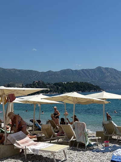 The image depicts a sunny day at a beach in Budva, Montenegro. The scene is filled with beachgoers relaxing on lounge chairs under large, light-colored umbrellas. The water is a clear turquoise, and the beach is covered in pebbles. In the background, majestic mountains rise, covered in lush green vegetation, with a small town nestled along the coastline. The sky is a bright, clear blue, suggesting pleasant weather and midday.
Several people are visible, some sunbathing, others sitting or preparing to enter the water. A few individuals are swimming or wading in the sea. The overall atmosphere appears relaxed and leisurely, indicative of a vacation or a day spent enjoying the coastal scenery. There are no discernible signs or text present in the image. The composition suggests a typical summer day at a popular tourist destination.