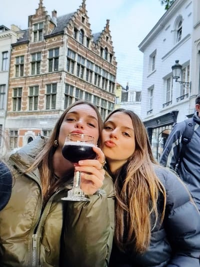 This image depicts a street scene in Forest - Vorst, Belgium. In the foreground, two young adult females are centrally positioned, looking towards the camera. The woman on the left, with long brown hair, wears an olive-green puffer jacket and holds a gold-rimmed stemmed glass containing a dark, foamy liquid. She has gold hoop earrings and a ring on her right hand. The woman on the right, also with long brown hair, wears a dark gray puffer jacket and a ring on her left hand. Both women have their lips pursed. The background features a European street under an overcast sky. A multi-story building constructed of reddish-brown brick with multiple windows and two distinct stepped gables is visible in the left and center background. To the right of this building stands a lighter-colored, possibly white, building with arched windows and a black wall-mounted lantern. A partially visible male figure, wearing a dark jacket and a backpack, is seen walking away on the far right of the frame.