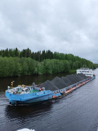 A barge, loaded with dark cargo, moves along a river. The barge is marked with the text "ОКСКИЙ 57". Lush green trees line the riverbanks, with a cloudy sky above.
The image was taken near the city of Depo, Russia.