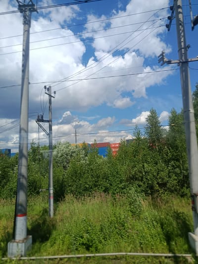 The image depicts an outdoor scene likely taken from a moving vehicle, showing a bright, cloudy sky with a predominantly blue hue. In the foreground and mid-ground, several utility poles with wires are visible, along with lush green foliage and tall grass. In the background, there are rows of shipping containers, some of which appear to be red and blue, and bear the inscription "CK LINE". The presence of shipping containers and the inscription suggests a commercial or industrial area, possibly a port or logistics hub. The overall environment suggests a sunny day in a semi-urban or industrial setting, characterized by vegetation and infrastructure related to transportation and logistics. The location is identified as Elektrougli, Russia.