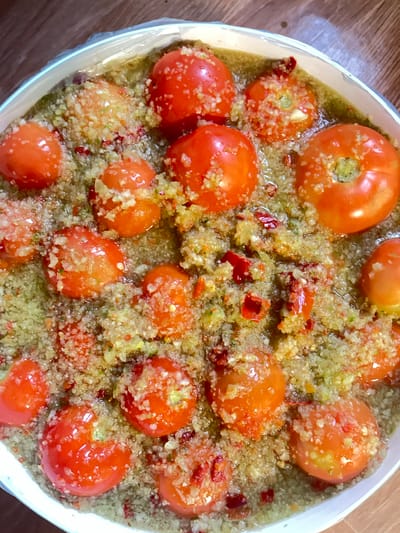 A close-up, overhead shot shows a white bowl filled with cherry tomatoes submerged in a pale green, chunky liquid. The tomatoes are whole and red, with some appearing slightly wrinkled. The liquid is textured with finely chopped ingredients, including small red pieces that appear to be chili peppers, and lighter colored, granular pieces that could be garlic or ginger. The bowl is placed on a wooden surface. There are no people visible in the image. The scene appears to be domestic and is located in Preobragenskaya, Russia.
