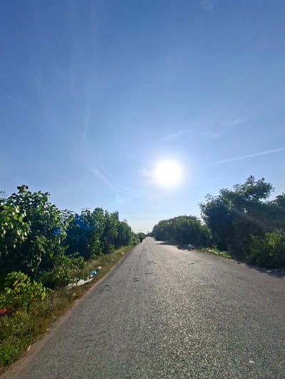 A long, empty asphalt road stretches into the distance under a clear blue sky with the sun shining brightly. Lush green trees line both sides of the road, with some scattered litter visible along the edges. A lone figure on a motorcycle is visible far down the road, traveling away from the camera. The scene appears to be in a rural or semi-rural area, possibly on the outskirts of Sumbokrayek, Indonesia, given the presence of dense vegetation and the overall undeveloped appearance of the roadside. The road surface is somewhat worn, with visible cracks and patches. The sunlight creates a strong glare on the asphalt.