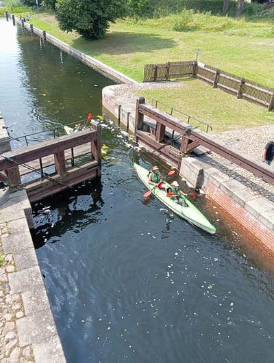 The image shows a canal lock with two kayaks navigating through it. In the foreground, a green and white kayak with two occupants wearing life vests and hats is being paddled. The occupant in the front is wearing a light-colored life vest and a hat, while the occupant in the back is wearing a green life vest and a dark hat. They are paddling with red-bladed paddles. Behind them, another yellow kayak with one person wearing a life vest and holding a red paddle is visible, partially obscured by the lock gate. The lock structure itself is made of wood and stone. The canal has dark water with some white foam and lily pads. The surrounding area features a grassy embankment with trees and a wooden fence. The scene is set in the city of Mikaszówka, Poland, a location known for its canals and locks connecting the Augustów Canal.