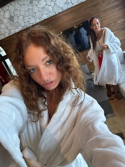 The image shows a selfie of a woman with curly hair wearing a white bathrobe. In the background, another person is also wearing a white bathrobe and lying on a surface. A dark table and a grey couch are visible in the background. The location appears to be indoors, possibly a hotel room or spa. The setting resembles modern interior design with elements such as wood paneling, stone flooring, and a flat-screen TV. There is no clear evidence to confirm that this photo was taken in Cat Cat Village, Vietnam.