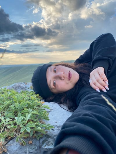 A woman lies on a rocky surface with green plants growing from the rocks. She is wearing a black hooded sweatshirt and a black beanie. Her head is turned towards the camera, and she is looking at the viewer. Her right hand is resting on her chest, and she has long, pointed fingernails painted white. In the background, there are rolling green hills under a cloudy sky. The location is the area around Абзаново, Russia.