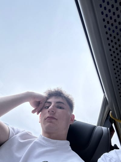 The scene depicts the interior of a vehicle, likely a bus or coach, located in Dobrodan, Bulgaria. A young male, appearing to be in his late teens or early twenties, with light brown, curly hair and a fair complexion, is centrally positioned and looking directly forward towards the camera. His left hand is resting on the left side of his forehead, fingers slightly curled. He is wearing a white t-shirt. To the far right, a portion of another individual's white t-shirt and shoulder is visible. The immediate environment includes a dark, possibly black, vehicle seat with a textured or stitched pattern, and an overhead grey panel with a perforated design along the top right edge. A thin window frame separates the interior from a bright, overcast sky visible through the upper portion of the vehicle's window. A yellow strap or handle is partially visible on the far right.