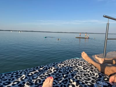 This image captures a sunny day at the Chiemsee lake in Gstadt am Chiemsee, Germany. In the foreground, a person's legs and feet, one with red nail polish, are resting on a leopard-print blanket on a wooden dock. To the right, another person's bare feet are visible. The lake extends into the distance, with several sailboats on the horizon. Further out, a wooden platform serves as a diving area for a group of people. Some individuals are swimming or wading in the water, and one person is on a small, green inflatable raft. The sky is a clear blue with a few wispy clouds. The scene suggests a leisurely summer day spent enjoying the water.