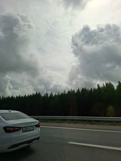 A white Lada Vesta sedan with a Russian license plate (M084HH 716) drives on a highway. The car is seen from the rear passenger side. The highway is bordered by a dense forest of dark green trees and a metal guardrail. The sky is heavily overcast with dramatic grey clouds, suggesting impending rain or a stormy weather condition. The image appears to be taken from another vehicle moving on the same highway, giving a dynamic perspective of travel. The overall scene suggests a journey through a natural, possibly remote, area. The location is identified as the city of Pokrovka, Russia.