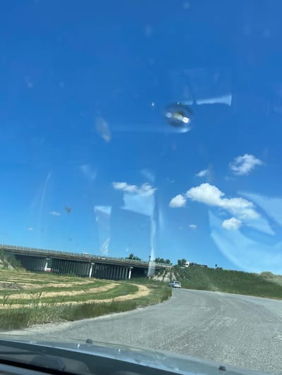 The image captures a view from a vehicle on a sunny day, looking towards a bridge and a rural landscape. The sky is a vibrant blue with scattered white clouds. The road ahead curves to the right, with a grassy embankment on the right side and a field with cut grass in a circular pattern on the left. A concrete bridge with multiple support pillars spans across the field and road. A white car is visible driving on the road ahead, and a white truck is parked on the embankment near the bridge. There are some reflections on the windshield, including a circular distortion with light reflecting off it, and some smudges or dust. The scene appears to be on the outskirts of Tatarka, Russia, given the context provided.