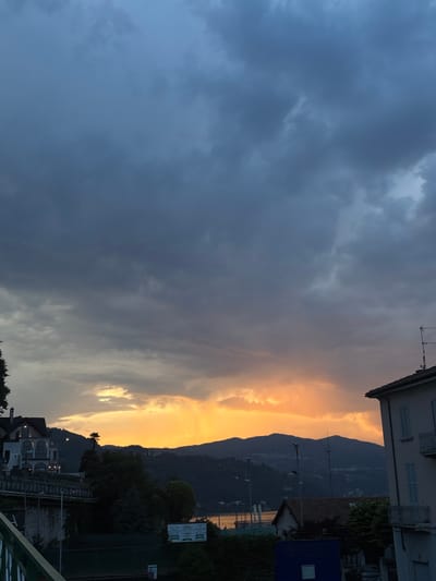 The image captures a twilight scene in Arona, Italy, looking out over Lake Maggiore. A dramatic sky dominates the upper portion of the frame, filled with dark, heavy clouds that are beginning to break to reveal a vivid orange glow of sunset or sunrise from the horizon. The light illuminates the underside of some clouds and casts a warm hue across the distant, silhouetted hills that line the far side of the lake.

In the foreground and midground, elements of the town of Arona are visible. On the left, a multi-story building with ornate architectural details, possibly a villa or hotel, sits on elevated ground. Its windows are dark, indicating either interior lighting or the fading light of day. Below this building, a retaining wall and some greenery are present, suggesting a tiered landscape. Further to the right, another building with a tiled roof and blue shutters is partially visible, its facade suggesting a residential or commercial structure. A television antenna is mounted on its roof.

The lake itself is visible in the middle ground, its surface reflecting some of the sky's colors, though it appears calm. In the distance, along the lakeshore, there are hints of other structures, possibly a promenade or smaller buildings. The overall atmosphere is one of dramatic natural beauty meeting the human presence of the lakeside town. There are no people clearly visible in the image.