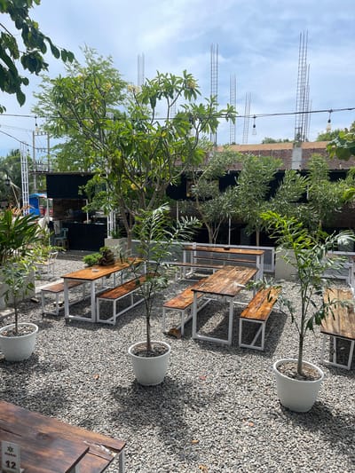 This image depicts an outdoor seating area at a cafe or restaurant in Pulokiton, Indonesia. The ground is covered with small, grey gravel. Several wooden picnic tables with white metal frames are arranged on the gravel. Some tables are empty, while others have benches that are unoccupied. Small trees and potted plants are interspersed throughout the seating area. In the background, a building under construction is visible, with exposed rebar and brickwork. String lights with visible bulbs hang above the seating area, and there are power lines and poles in the distance. A black corrugated metal structure is part of the building's facade. The sky is partly cloudy. There are no people or animals clearly visible in the frame.