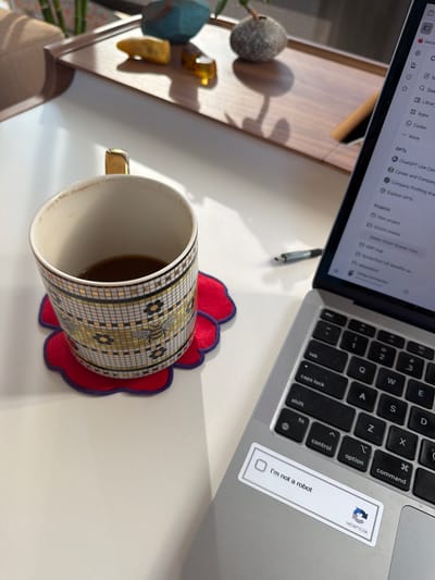 The image shows a close-up, overhead view of a laptop, a mug, and some decorative objects on a white desk. The laptop is open, displaying a screen with various icons and text, including "GPTS", "ChatGPT Use Cases", and "Explore GPTS". A "reCAPTCHA - I'm not a robot" sticker is visible on the laptop's palm rest. The mug has a mosaic-like pattern with bees and flowers, and it contains a dark liquid, likely coffee. A red, flower-shaped coaster sits beneath the mug. To the left of the mug are two abstract decorative objects, one teal and one gold, casting shadows on the desk. The lighting suggests it is daytime, with sunlight creating distinct shadows. The scene appears to be a home office or workspace, given the presence of a laptop and personal decorative items. No people are visible in the image.