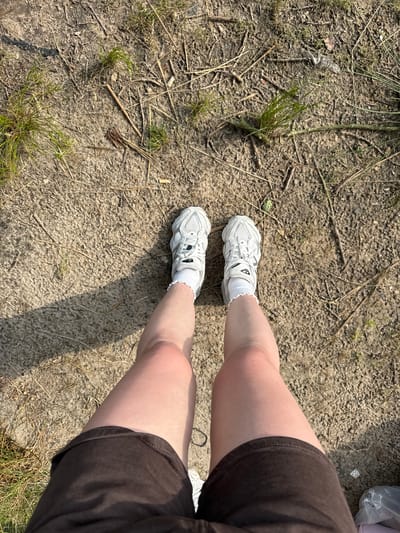 A person stands on a dirt path with scattered twigs and dry grass. The perspective is a top-down view of their legs and feet. They are wearing dark brown shorts, white socks with a ruffled edge, and white sneakers with grey accents. The sneakers appear to be athletic shoes. The ground is a mix of dry soil, small pebbles, and sparse patches of green grass. Sunlight casts a shadow from the person onto the ground to their left. There is no identifiable urban or natural landmark within the image to indicate a specific location within Turnhout, Belgium.