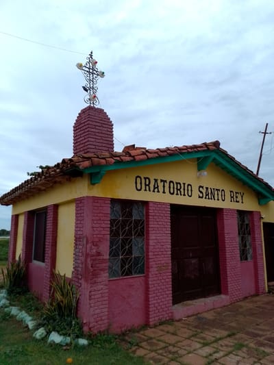 An exterior shot shows a small, colorful religious building identified as "ORATORIO SANTO REY" (Holy King Oratory), located in Concepción, Paraguay. The building features vibrant yellow walls with sections of reddish-pink brickwork and trim. The roof is made of terracotta tiles, with a bright turquoise fascia board under the eaves, some of which appear to have small plants growing on them.

A prominent red brick chimney or tower rises from the roof, topped with an ornate wrought-iron cross. This cross is decorated with several colorful, spherical ornaments, possibly lights or baubles, in red, yellow, green, and white.

The main facade has a dark brown double door at the center, flanked by two windows with decorative metal grates. Another window is visible on the left side of the building. The ground immediately around the building is a mix of green grass and square paving stones. Along the base of the left side, there's a small garden bed with spiky green and yellow-leafed plants, bordered by light-colored rocks. A single orange object, possibly a fruit, lies on the grass in the foreground.

The sky is overcast with soft, diffused light, suggesting either early morning, late afternoon, or a heavily cloudy day. No people are visible, and there is no apparent activity taking place, conveying a sense of quietude. A thin wire crosses the upper left portion of the sky, and a utility pole with some wiring is partially visible on the right side behind the building.
