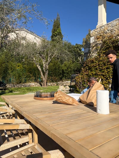 This image captures an outdoor scene in a garden setting in Mougins, France, under clear blue skies and bright sunlight. A large, light-toned wooden slatted outdoor dining table occupies the foreground and midground, with several matching wooden chairs featuring woven rope seats and backs partially visible.

On the table, a circular wooden tray holds six dark-tinted drinking glasses. Adjacent to these are two brown paper bags, one standing upright and partially open, and a white rectangular box, indicating packaged items. A white roll of paper towels stands vertically on the table's right side.

Two individuals are positioned around the table. To the right, a young male with short, dark, curly hair, dressed in a black long-sleeved top and blue shorts, stands, looking downwards towards the table. His left hand holds a clear glass jar. Partially obscured by the upright brown paper bag, a younger individual with braided hair, wearing a light blue top, appears to be seated or kneeling, engaged with the contents of the bag or box.

The background features a green lawn bordered by a low stone wall and a dense green bamboo hedge. A mature olive tree with sparse foliage stands centrally in the midground. To the right, a stone wall covered in climbing ivy rises, with a section of a light-colored stucco building and a prominent column visible above it. In the further background, a pale building with a tiled roof is partially visible through tree branches. A dark barbecue grill is situated on the lawn to the left of the stone wall. A tall, slender cypress tree is visible on the right side of the frame.