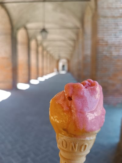 A close-up of a two-tone ice cream cone is centered in the foreground against a blurred architectural background. The ice cream is orange on the bottom and pink on the top. The cone itself has “CONO” embossed into its surface.
The background shows a long arcade of arched walkways, characteristic of Italian architecture. The arcade recedes into the distance, where a brighter doorway is visible. The location is likely in Mezzana Sant'Antonio, Italy, based on the prompt. The floor of the arcade is paved, and sunlight is visible in patches along the arcade floor. The perspective is slightly low, suggesting the photo was taken by someone holding the ice cream at chest level. There is no evidence of people in the immediate surrounding.
