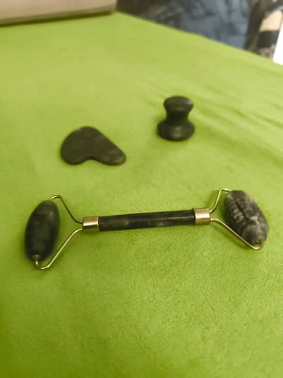 The image shows a close-up, eye-level shot of a jade facial roller and two gua sha tools arranged on a bright green, textured surface, likely a cloth. The main subject is a double-ended jade roller, positioned horizontally across the lower half of the frame. The roller has a smooth, dark jade large ball at the left end and a smaller, ribbed jade roller at the right end. Both are attached to a dark jade handle with thin, gold-toned metal wire supports. To the left and slightly behind the roller is a dark, heart-shaped gua sha tool. Further back and to the right is another dark, mushroom-shaped gua sha tool. The background is softly blurred, showing hints of a light-colored surface above and a darker, possibly blue, textured surface behind that, suggesting an indoor setting. There is no visible text in the image. The lighting appears even, with no strong shadows, and the time of day and weather are not discernible. The overall impression is one of a skincare or beauty product laid out for use, with a focus on the tools themselves. No people are visible in the frame.