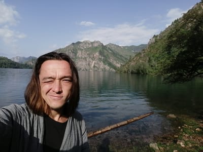 In Yaryktash, Kyrgyzstan, a man with long, dark hair is taking a selfie by a clear lake. He is wearing a grey, patterned open robe over a black t-shirt, and he is winking. The lake's surface is calm, reflecting the blue sky and scattered white clouds. In the background, imposing, rugged mountains rise steeply from the opposite shore, their slopes covered with a mix of rocky scree and verdant green vegetation. The sun casts shadows across the man's face and the foreground. A fallen log lies partially submerged in the shallow water near the shore, which is composed of grass and scattered stones. The overall scene depicts a serene natural landscape.