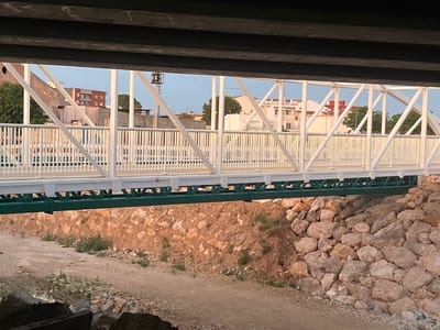 The image depicts a view from beneath an overhead structure, looking through and beyond a white metal pedestrian bridge in Alcàsser, Spain. Dark, horizontal beams of the unseen overhead structure frame the top of the image. The prominent midground feature is a white-painted metal truss bridge, characterized by a crisscross pattern of support beams and uniform vertical railings. A substantial dark green metal beam runs horizontally beneath the white bridge deck, forming its primary support structure.

Below the bridge, the ground consists of a light brown dirt path with scattered rocks and sparse green vegetation. To the left, a sloped bank of reddish-brown earth is interspersed with numerous light-colored stones and small patches of green foliage. To the right, a dense arrangement of large, irregular, light-colored boulders forms a steep embankment or retaining wall.

Through the white framework of the pedestrian bridge, a semi-urban background is visible under a pale blue sky. Various buildings are present: a multi-story, reddish-brown building with white-framed windows on the left, and numerous lighter-colored buildings, some with red or terracotta-tiled roofs, extending across the frame. Green trees are scattered throughout the background, partially obscuring some structures. On the far left, a tall, slender utility pole or lattice tower stands prominently. The scene is illuminated by soft, warm light, suggesting either late afternoon or early morning. No individuals or active subjects are visible in the frame.