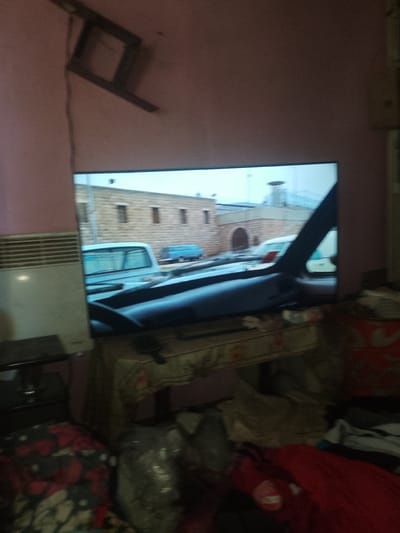 The image depicts an indoor scene, likely a living room, in Rus'ka Dolina, Ukraine. A large television screen is prominently displayed, showing an outdoor scene with buildings and vehicles. To the left of the television, a white radiator is visible against a pink wall. The foreground shows a cluttered surface with various objects, including what appear to be blankets and bags. A dark furniture piece is partially visible beneath the radiator. The overall impression is of a lived-in space with a focus on entertainment.