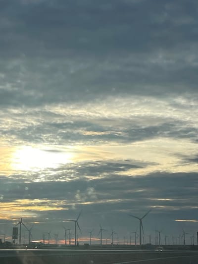 The image depicts a large wind farm in Tébar, Spain, under a predominantly cloudy sky, consistent with either sunrise or sunset. The upper two-thirds of the frame are occupied by layered grey and broken lighter clouds. An intense, partially obscured light source, likely the sun, is positioned on the far left, casting a yellow-orange glow across the lower cloud layers.

Numerous wind turbines, silhouetted darkly against the brighter horizon, span the mid-ground and extend into the distance, indicating an expansive installation. Some turbine blades exhibit motion blur, suggesting rotation. In the foreground, a dark, multi-lane highway or road traverses horizontally across the bottom of the image. Two vehicles with illuminated headlights are visible on the road surface: one on the left in the mid-distance and another on the right, closer to the viewer. A dark, rectangular structure, possibly a road sign, stands on the left side of the highway, in front of the turbines. No individuals are discernible.