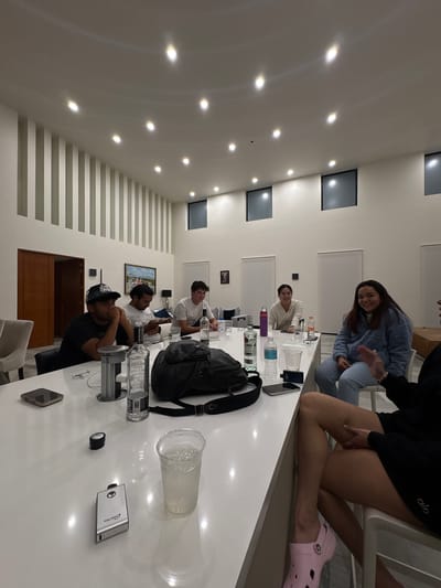 **Location:** Santiago Etla, Mexico (based on limited information, cannot be definitively confirmed).
**Scene:** A well-lit, modern interior room. A long, white table dominates the scene, occupied by multiple individuals.
**People:** At least six people are partially or fully visible.
* One woman, seated on the table's edge, wears a black outfit and pink Crocs.
* Another woman is seated at the table in blue pajamas.
* Several men are seated around the table.
**Objects:** The table is covered with items, including:
* A black backpack.
* A large plastic cup.
* Bottles (some possibly alcohol).
* Electronic devices (phone, possibly a camera).
* Various other containers.
**Actions/Interactions:** The people appear to be socializing or possibly participating in a casual gathering. Some individuals are looking at the camera.
**Environment:** The room has white walls with recessed lighting fixtures. One wall features horizontal indentations. Artwork and a door are visible in the background.