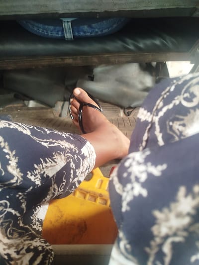 The image shows a person's right foot wearing a black flip-flop. The foot is resting on a textured surface, possibly the floor of a vehicle, which appears to be a tricycle or auto-rickshaw given the presence of a yellow plastic component visible beneath the foot. The person is wearing dark blue pants with a white floral pattern. In the background, a dark seat and a blue denim material are visible, suggesting an indoor or covered seating area. The lighting is moderate, and there are no clear indicators of the time of day or weather conditions. The image is taken from a low angle, focusing on the person's lower leg and foot. The overall impression is a candid shot, possibly taken while traveling in a vehicle in Uselu, Nigeria. No text is visible in the image.
