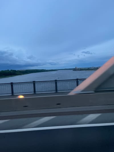 The image shows a view from a bridge over a body of water, likely a river, under a cloudy sky. In the distance, across the water, is a shoreline with trees and some structures, possibly buildings. There is a railing along the side of the bridge in the foreground, and part of the bridge structure is also visible. A bright orange light is visible on the side of the bridge. The water appears choppy, suggesting wind. The sky is overcast with dramatic clouds. The scene is captured during what appears to be dusk or early evening. There are no people visible in the image. The location is identified as the city of Il'inskoe (Lomovskaya S/A), Russia.