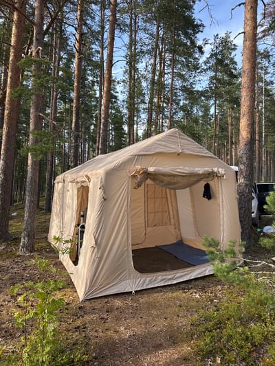 The image shows a large, tan-colored canvas tent set up in a pine forest. The tent is open, revealing a dark blue sleeping mat inside. A black garment is hanging on a loop on the exterior of the tent. The ground is covered with pine needles, moss, and small green plants. Tall pine trees surround the tent, with the sky visible through their branches. A white SUV with its doors open is parked behind the tent, to the right. The scene suggests a camping trip in a wooded area near Priladozhskoe, Russia.