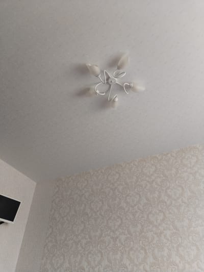 The image shows the interior of a room, likely a bedroom or living room, featuring a ceiling with a white, star-shaped light fixture and patterned wallpaper. A corner of a black television screen is visible on the left side of the image. The lighting is soft and diffused. The room appears to be located in Troitskoye, Russia, although no specific landmarks or identifying features are present in the image to confirm this location. There are no people or animals in the frame.
