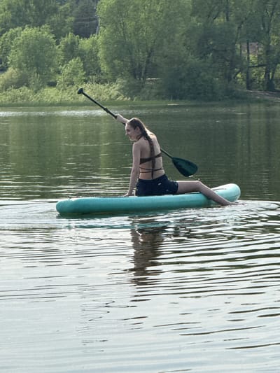 A woman is on a light blue paddle board in the water. She is leaning over, placing a black paddle into the water. The surrounding water is slightly disturbed with reflections. Trees are visible on the bank to the left. The scene appears to be Novosinkovo, Russia.