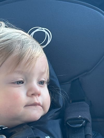 The image displays a close-up, rotated view of a young child, likely a baby or toddler, lying in a car seat or stroller. The child's head occupies the lower-left portion of the frame, with their body extending towards the upper-right.

The child has light blonde or platinum-colored hair and striking blue or grey eyes, which are open and looking towards the top-right of the image. Their skin appears fair with a natural blush on the cheeks, and their mouth is slightly agape or in a neutral, relaxed position.

The child is situated against a dark blue, finely textured fabric, which is characteristic of the upholstery found in child car seats or strollers. In the upper-left area of the frame, near the child's head, a distinctive white logo is visible, composed of several overlapping circular rings. This logo identifies the brand as Bugaboo. To the right of the child's head, a black, padded harness strap with a shiny metal D-ring or buckle component is visible, indicating a safety restraint system. The overall lighting suggests it is daytime, casting a soft, even illumination on the scene. The child appears calm and observant.