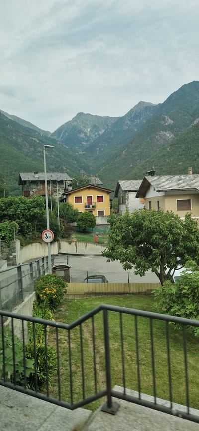 This is a landscape photograph taken outdoors, likely in a mountainous region given the prominent mountains in the background. The scene depicts a small village with several houses nestled on a hillside. The houses appear to be traditional in style, with pitched roofs and balconies. There's a yellow house with red window frames and balconies, and other houses with grey tiled roofs. Lush green trees and vegetation surround the buildings, suggesting a rural or semi-rural setting.
A road or pathway is visible, with a parked car. A speed limit sign with "3,50 t" is also present, indicating a weight restriction. The sky is overcast, suggesting a cloudy day. The overall atmosphere is peaceful and serene. The orientation of the image is portrait, with the top of the frame showing the sky and mountains, and the bottom showing the foreground with a railing and some foliage. The lighting suggests it is daytime, but not necessarily bright sunlight. The location is identified as Challand-Saint-Victor, Italy. There are no people visible in the frame, and the focus is on the natural scenery and the architecture of the village.