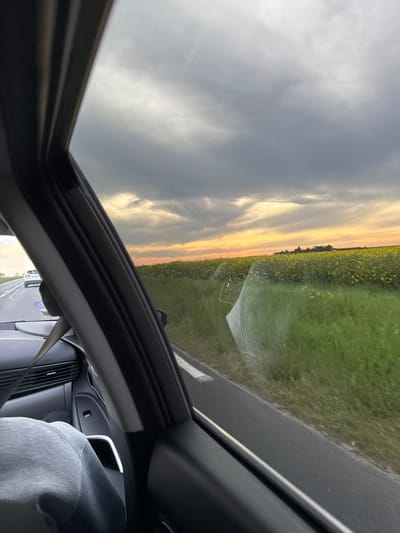The image is a view through the open sunroof of a car in Gometz-la-Ville, France. Outside, the landscape consists of a grassy field alongside a road with a view of the sky above. The sky has cloudy conditions with some yellow in the horizon, suggesting it might be either sunrise or sunset.
Inside the car, the interior is visible, including parts of the dashboard, air vents, and a passenger wearing a gray garment in the passenger seat. A safety belt is also seen in the foreground.