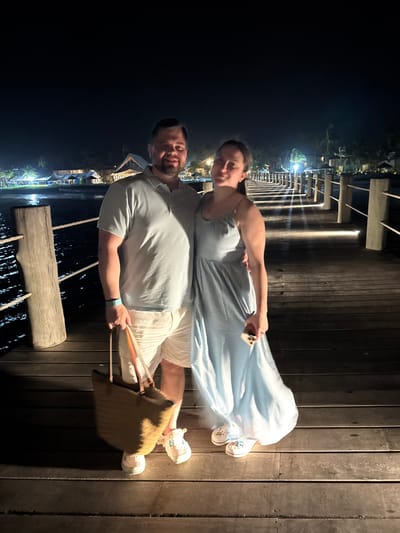 A man and woman pose at night on a wooden pier. The woman, in a light blue dress, holds a phone. The man wears a light blue shirt and white shorts, holding a straw tote bag. They stand on a wooden boardwalk with lights visible in the distance. A rope railing separates them from the dark water below. The photo is taken at an angle, with the pier extending towards the background.