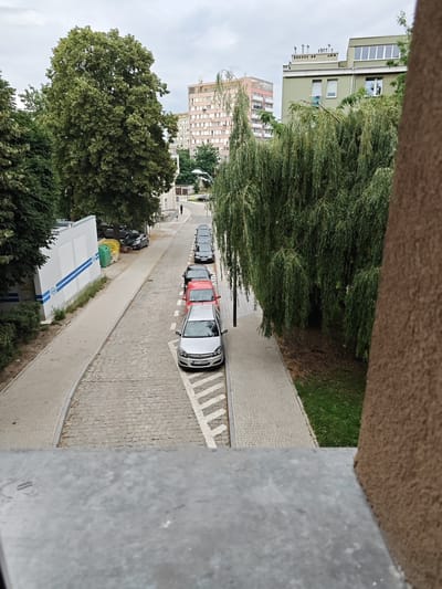 The image shows a street in the city of Stargard, Poland, with several cars parked along the side. The street is paved with cobblestones, and there is a sidewalk on the left side of the image. There are also several trees and buildings visible in the background. The sky is overcast. A single person is visible walking down the street in the distance. The foreground is occupied by a concrete surface, possibly a ledge or railing. The cars are lined up facing away from the camera, with a silver car closest to the foreground.