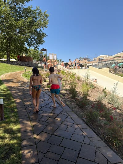 The image depicts a sunny day at a water park in Cavriglia, Italy. In the foreground, a young girl and boy walk away from the camera on a paved pathway. The girl wears a bikini with a patterned bottom and the boy wears red and blue swim trunks. To the right of the pathway, a water slide curves down a grassy slope, with a child in the process of sliding down. In the background, several adults and children are visible near the top of the water slide and around a wooden hot tub. The environment includes lush green trees, manicured lawns, and what appear to be buildings with terracotta roofs in the distance. The sky is a clear, bright blue.