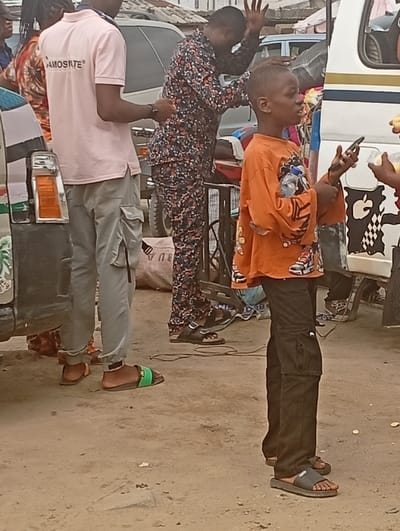 The image captures an outdoor scene in Igwuruta, Nigeria, featuring multiple individuals and vehicles on dusty ground. In the foreground, a young male, appearing to be a child or pre-teen, stands facing left. He wears an oversized orange long-sleeved shirt with graphic designs, dark cargo pants, and dark slides. He holds a black smartphone in his left hand and extends his right hand to receive light-colored food items in clear plastic containers from an unseen person to his right.
To the young male's left, a man stands, partially obscured by a light-colored vehicle on the far left. This man wears a light pink polo shirt with "AMOSITE" printed on the chest, light grey cargo pants, and green and black slides. His right hand is extended forward. Behind him and slightly to the right, another man, dressed in a dark, multi-patterned long-sleeved shirt and matching pants, stands with his head bowed and both hands raised above his head, palms open.
Vehicles include a partially visible white van or bus on the right with a black and white checkered pattern and a visible "Apple" logo-like sticker. Blurred individuals, background structures, and dark electrical wires on the ground near the feet of the man in patterned attire are also present, suggesting a public or market environment.
